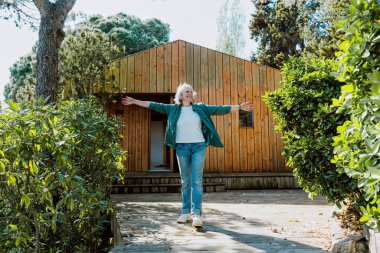 Mutlu son sınıf öğrencisi kadın kollarını açmış, özgürlüğünü ve doğayla bağlantısını deneyimliyor modern bir kulubenin önünde, yemyeşil ağaçlar ve çalılarla çevrili.