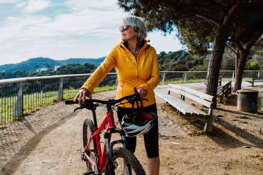 Dağ bisikletinin yanında duran yaşlı kadın, sarı bir ceket ve güneş gözlüğü takıyor, dağ yolunda temiz havanın ve manzaranın tadını çıkarıyor, emeklilikteki sağlık ve özgürlüğün somutlaşmış hali.