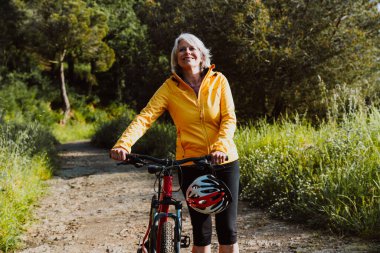 Kirli bir yolda dağ bisikletinin yanında duran, gülümseyen ve mesafeye bakan, manzaralı yeşil bir dağ zeminiyle aktif ve sağlıklı bir yaşam tarzını kucaklayan yaşlı bir kadın.