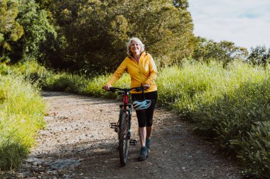 Kirli bir yolda dağ bisikletinin yanında duran, gülümseyen ve mesafeye bakan, manzaralı yeşil bir dağ zeminiyle aktif ve sağlıklı bir yaşam tarzını kucaklayan yaşlı bir kadın.