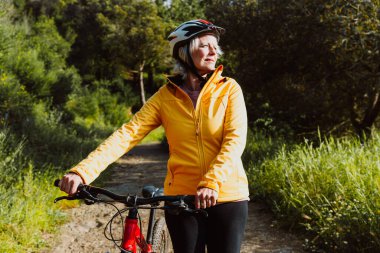 Kirli bir yolda dağ bisikletinin yanında duran, gülümseyen ve mesafeye bakan, manzaralı yeşil bir dağ zeminiyle aktif ve sağlıklı bir yaşam tarzını kucaklayan yaşlı bir kadın.