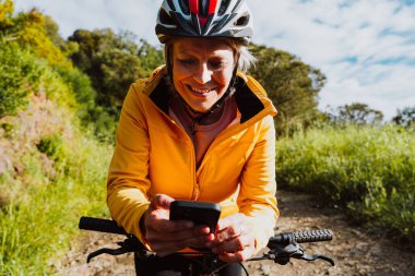 Kasklı ve sarı ceketli, gülümseyen, akıllı telefonunu kontrol etmek için bisikletini durduran, sağlıklı spor salonunun ve modern teknolojinin tadını çıkaran son sınıf öğrencisi.