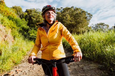 Kirli bir yolda dağ bisikletinin yanında duran, gülümseyen ve mesafeye bakan, manzaralı yeşil bir dağ zeminiyle aktif ve sağlıklı bir yaşam tarzını kucaklayan yaşlı bir kadın.