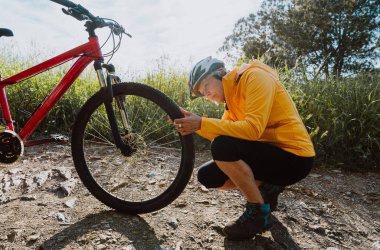 Bisiklet giysisi giymiş yaşlı bir kadın toprak bir patikada çömelmiş kırmızı dağ bisikletinin ön lastiğini dikkatle inceliyor, bakım ve aktif yaşam tarzını gösteriyor.