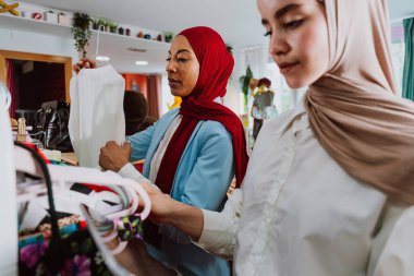 Başörtüsü takan Müslüman kadınlar askılara bakıyorlar, çeşitli moda mağazalarında modern bir alışveriş deneyimi yaşarken nesneleri dikkatlice seçiyorlar.