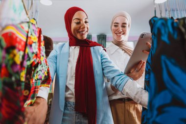 İki modern Müslüman kadın moda seçeneklerini tartışıyor, biri dijital tablet tutarken askılardaki kıyafetlere bakıyor, modern bir perakende pazarlama ortamında alışveriş ve stili gösteriyor.
