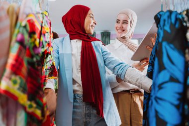 İki modern Müslüman kadın moda seçeneklerini tartışıyor, biri dijital tablet tutarken askılardaki kıyafetlere bakıyor, modern bir perakende pazarlama ortamında alışveriş ve stili gösteriyor.