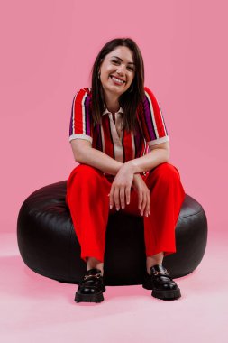 Genç, gülümseyen bir kadın rahat ve neşe dolu siyah bir koltukta oturuyor, parlak çizgili bir polo tişört ve pembe bir arka planda kırmızı geniş paçalı pantolon giyiyor.