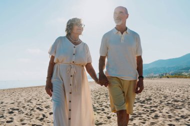 Güneşli bir sahilde el ele tutuşan ve yalınayak yürüyen yaşlı bir çift, emekliliğin tadını çıkarıyor ve deniz kenarında rahatlatıcı bir yaz tatili, mutluluk ve beraberliği ifade ediyor.