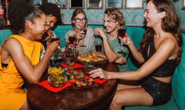 Çeşitli genç yetişkinler sosyal bir toplantının keyfini çıkarıyor, gülümsüyor, gülüyor ve bir bar veya restoran masasında ferahlatıcı içecekler ve aperatiflerle etkileşime geçiyorlar.