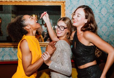 Üç farklı genç kadın parti elbiseleri giyip birlikte poz vererek, kokteyller ve atıştırmalıklarla şık bir bar veya salon ortamında özel kutlamalar yapıyorlar.