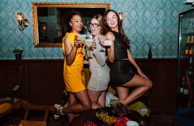Üç farklı genç kadın parti elbiseleri giyip birlikte poz vererek, kokteyller ve atıştırmalıklarla şık bir bar veya salon ortamında özel kutlamalar yapıyorlar.