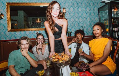 Arkadaşlar şampanya kadehlerini tokuştururken bir kadın şık bir barda, neşeli bir akşam kutlaması sırasında aperatifler sunuyor.