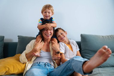 Asyalı bir aile evde, gülümseyen Asyalı bir anne evde iki oğluyla koltukta oturuyor, bir çocuk da omuzlarında oturuyor. Aile ve ebeveynlik kavramı