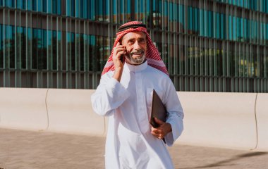 Yetişkin bir orta doğulu işadamı, bir dosya taşırken cep telefonuyla iletişim kuruyor, şehir caddesinde modern binaların yanında duruyor, küresel ticaret ve bağlantıyı temsil ediyor.