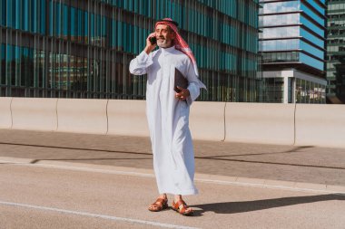 Yetişkin bir orta doğulu işadamı, bir dosya taşırken cep telefonuyla iletişim kuruyor, şehir caddesinde modern binaların yanında duruyor, küresel ticaret ve bağlantıyı temsil ediyor.