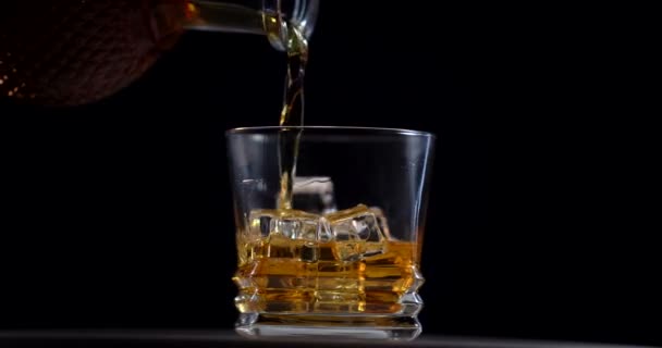 Whisky dorado vertiendo en el vaso de la botella. Con hielo cubos. El camarero vierte whisky de un decantador en un vaso. Alcohol. El vidrio gira. Fondo oscuro. — Vídeo de stock