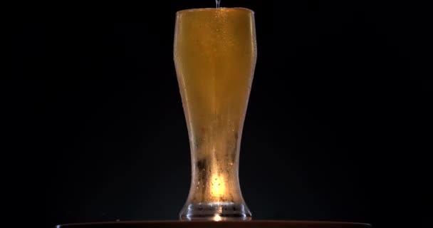 Холодное пиво в стекле вращается на поверхности с черным фоном. Детальный снимок пивных пузырьков в стекле. холодное пенное пиво наливая и брызги в стеклянную чашку на коричневом фоне — стоковое видео