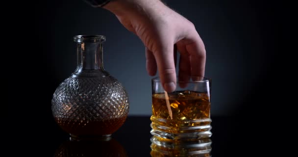 Uísque. O homem branco toma um copo com álcool escocês. Uísque na mesa refletida de vidro. Garrafa com uísque para trás. Fundo preto com luz branca. Conceito de férias — Vídeo de Stock