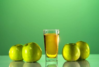 Yeşil elmalar bir yansıma ile masanın üzerinde uzanır. Onun yanında bir bardak elma suyu var. Yeşil arkaplan