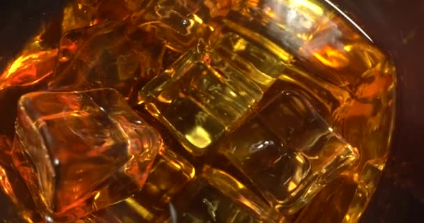 Ett glas whisky med iskub. Alkohol häller i glaset från flaskan. Whisky på klipporna. Slider skjuten. Mörk, rustik, bakgrund. Makroskott — Stockvideo