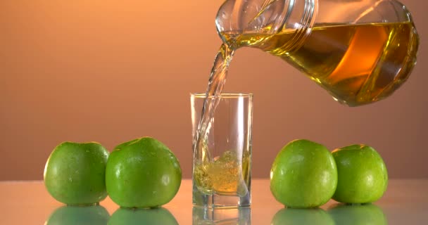 Äppeljuice i glas på det vita bordet Gröna äpplen. Göra färsk juice. Slow motion detaljerad bild av hälla läckra äppeljuice i ett transparent glas mot vit bakgrund. Närbild — Stockvideo