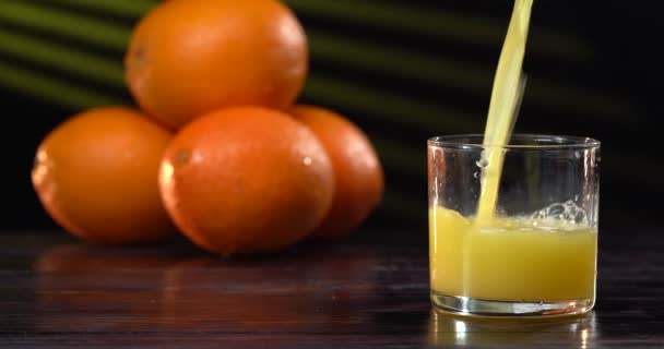 Sok pomarańczowy wlewa się do szklanki. Na stole w pobliżu są świeże pomarańcze. Ciemny stół. Ciekawe tło — Wideo stockowe