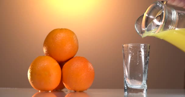 O suco de laranja derrama-se em um copo. há laranjas frescas na mesa nas proximidades. por do sol fundo — Vídeo de Stock