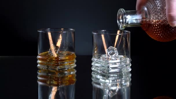 Gouden whisky gieten in het glas van de fles. Met ijsblokjes. Twee glazen op tafel. Reflecteer het oppervlak. Alcohol concept. — Stockvideo