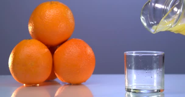 Verter el jugo de naranja en el vaso en fondo azul. Superficie reflejada. Naranjas frescas están sobre la mesa — Vídeo de stock
