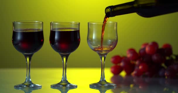 Verter vino tinto en una copa de botella oscura sobre el fondo amarillo. Tres vasos en superficie reflectante. Un racimo de uvas en la mesa. Alcohol — Vídeo de stock