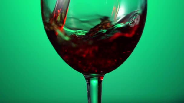 A vörösbor gyönyörű hullámot alkot. Bor öntés borospohárban zöld háttérrel. Közelről lőtték le. A vörösbor palackból serlegbe öntésének lassú mozdulata. Alacsony kulcs — Stock videók