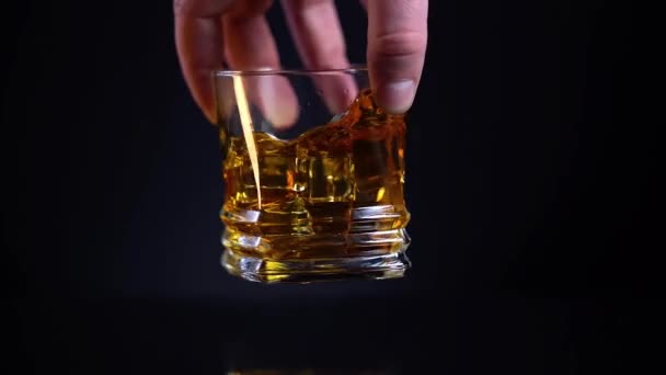 Male hand shaking a glass with tasty elegant Brandy or Cognac before trying it, super close-up. glass with cognac and shaking, alcoholic beverage. Luxury drink isolated on black background. 4k — Stock Video