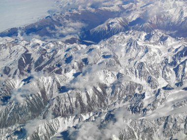 Bulutların altında gri karlı dağlar ve mavi gökyüzü. Uçak penceresinden bak. Seyahat kavramı