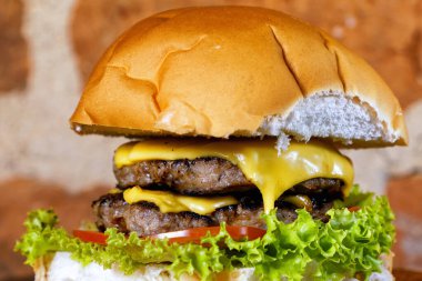 Pastırmalı, sarı peynirli, domatesli, yeşil salatalı, ahşap tahtanın altında arka planda kırsal duvarlı, nefis el yapımı çift katlı hamburger sandviçi.