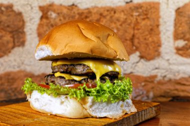 Pastırmalı, sarı peynirli, domatesli, yeşil salatalı, ahşap tahtanın altında arka planda kırsal duvarlı, nefis el yapımı çift katlı hamburger sandviçi.