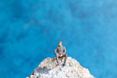 Minyatür insanlar. İş adamı mavi arka planda kayanın üzerinde oturuyor.