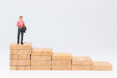 Yükselen tahta blokların tepesinde duran minyatür işadamı, başarıyı, kariyer büyümesini ve hedeflere ulaşmayı simgeliyor.