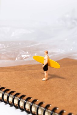 Küçük sörfçü figürü, yaratıcı bir konsept sahnesinde, çevresel sorunları, kirliliği ve okyanus farkındalığını sembolize eden plastik bir yaprağın yanında bir defter üzerinde duruyor..
