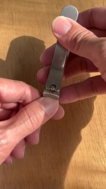 Makro yakın plan elleri paslanmaz çelik tırnak makasıyla tutarken doğal güneş ışığı altında tahta bir masada tırnaklarını kesiyor..