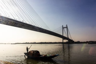 Kolkata, Batı Bengal, Hindistan - Ocak 2018: Kolkata şehrindeki Hooghly nehri üzerindeki ikonik Howrah köprüsünün altında tahta bir tekne kullanan bir adamın silueti.