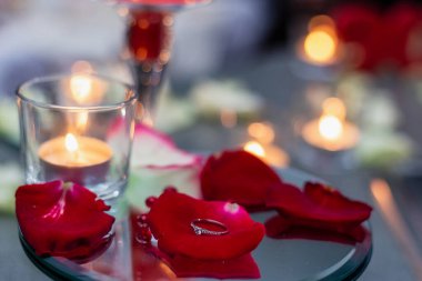 Alyans, mumların arkasındaki kırmızı gül yapraklarının üzerinde duruyor. Evlilik teklifi. Romantik atmosfer