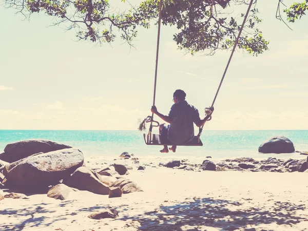 उष्णकटिबंधीय ग्रीष्मकालीन समुद्र तट पर एक स्विंग में युवा आदमी — स्टॉक फ़ोटो, इमेज