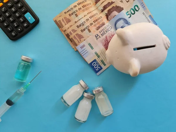 Meksika parası, domuz kumbarası, hesap makinesi, ilaç şişeleri ve mavi arka planda şırınga.
