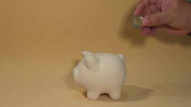 Bir adamın bej arka planda bir domuz kumbarasına bozuk para ve 5 euro yatırması.