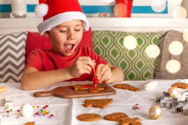 Noel Baba şapkalı mutlu bir çocuk mutfakta zencefilli kurabiye süslüyor. Noel ışıkları. Yüksek kalite fotoğraf