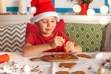 Noel Baba şapkalı mutlu bir çocuk mutfakta zencefilli kurabiye süslüyor. Noel ışıkları. Yüksek kalite fotoğraf