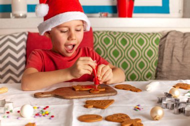 Noel Baba şapkalı mutlu bir çocuk mutfakta zencefilli kurabiye süslüyor. Yüksek kalite fotoğraf
