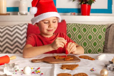 Noel Baba şapkalı mutlu bir çocuk mutfakta zencefilli kurabiye süslüyor. Noel ışıkları. Yüksek kalite fotoğraf