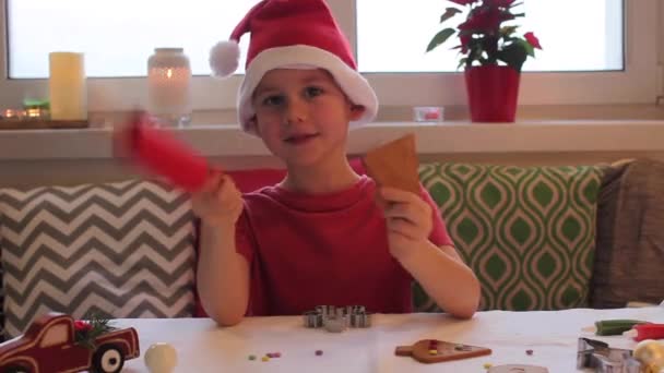 サンタクロースの帽子の幸せな男の子は台所で生姜クッキーを飾る。クリスマスライト. — ストック動画